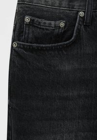 Gros plan sur la poche d'un jean en denim noir avec des coutures visibles et des rivets en métal sur la braguette à boutons et les bords de la poche.