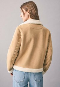 Beige Schaffelljacke mit strukturiertem Kragen, weicher Textur und sichtbaren Nähten, kombiniert mit hochgeschnittenen blauen Jeans.
