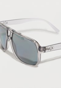 Γυαλιά ηλίου Armani Exchange με διάφανο πλαίσιο και φακούς με γαλάζια απόχρωση, με το λογότυπο της μάρκας στο πλάι, αποτυπωμένα σε λευκό φόντο.