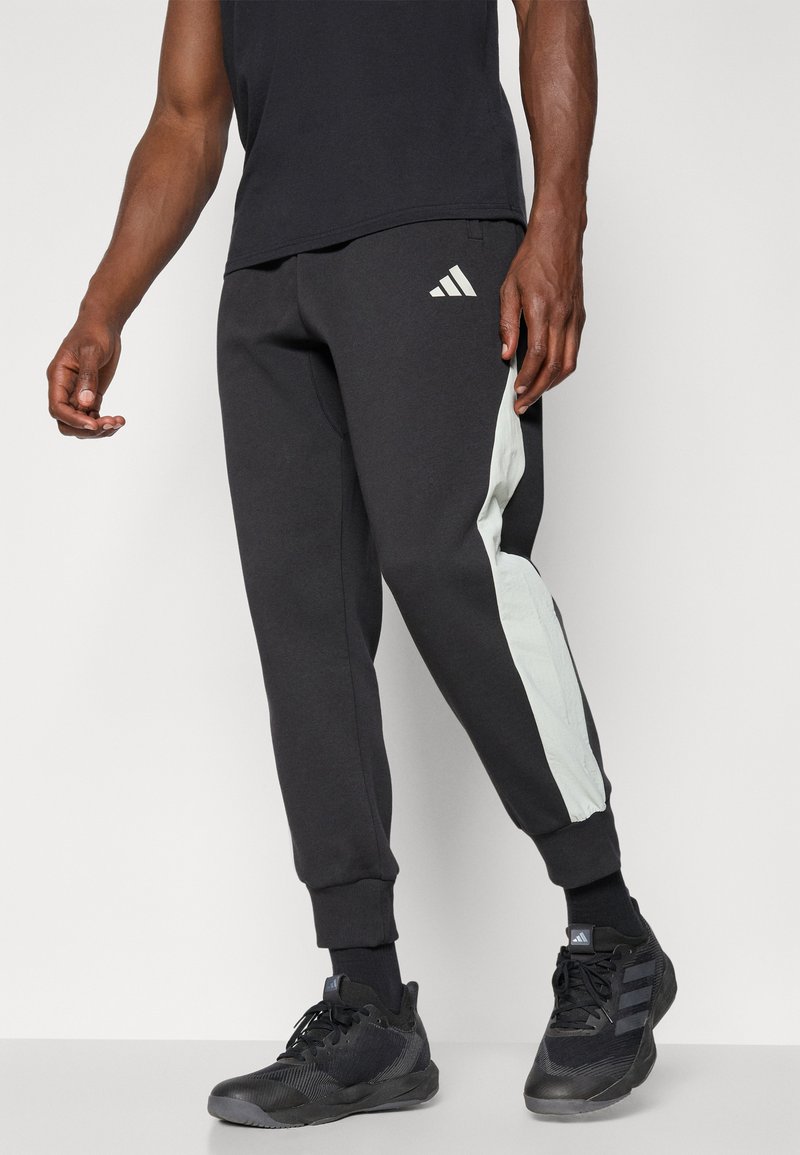 Mężczyzna w czarnych dresowych spodniach Adidas z białymi paskami po bokach, w czarnych skarpetach i czarnych sneakerach Adidas, stojący na jednolitym jasnym tle.