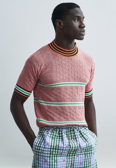 Pull en maille à col rond rouge avec motif texturé, rayures vertes et blanches, et détails en côtes noir-orange ; assorti à un short multicolore à carreaux.