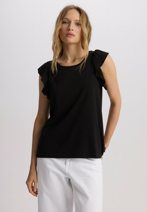 Czarna bluzka z krótkim rękawem z falbaniastymi akcentami na ramionach, wykonana z miękkiego materiału, w połączeniu z białymi spodniami. Prosty okrągły dekolt.