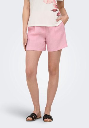 Femme portant un short rose, une chemise blanche à motif floral et des sandales noires à enfiler, debout avec une main dans la poche.