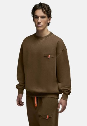 Jeune homme portant un sweat-shirt marron oversize et un pantalon avec un petit détail orange sur la poche poitrine et les cordons, sur un fond uni.