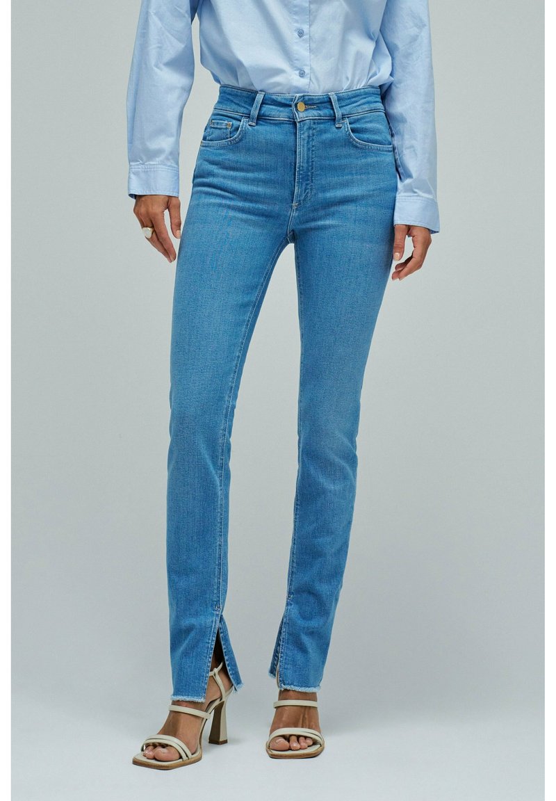 Frau trägt hellblaue Jeans mit vorderen Knöchelschlitzen, hellblaues Hemd mit Knöpfen und beige Riemchen-Sandaletten mit hohen Absätzen.