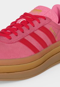 Ροζ σουέτ αθλητικό Adidas Gazelle με κόκκινες ρίγες, κόκκινα κορδόνια, κίτρινη λεπτομέρεια με λογότυπο και χοντρή σόλα από καουτσούκ.