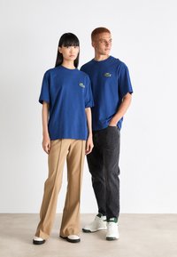 Camisetas de algodón azules con mangas cortas, con un logo bordado en verde; combinadas con pantalones beige y gris oscuro y zapatillas blancas.