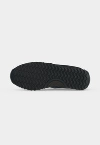 Semelle d'une chaussure noire avec un motif de bande de roulement ondulé et un talon arrondi, affichée sur un fond gris clair uni.