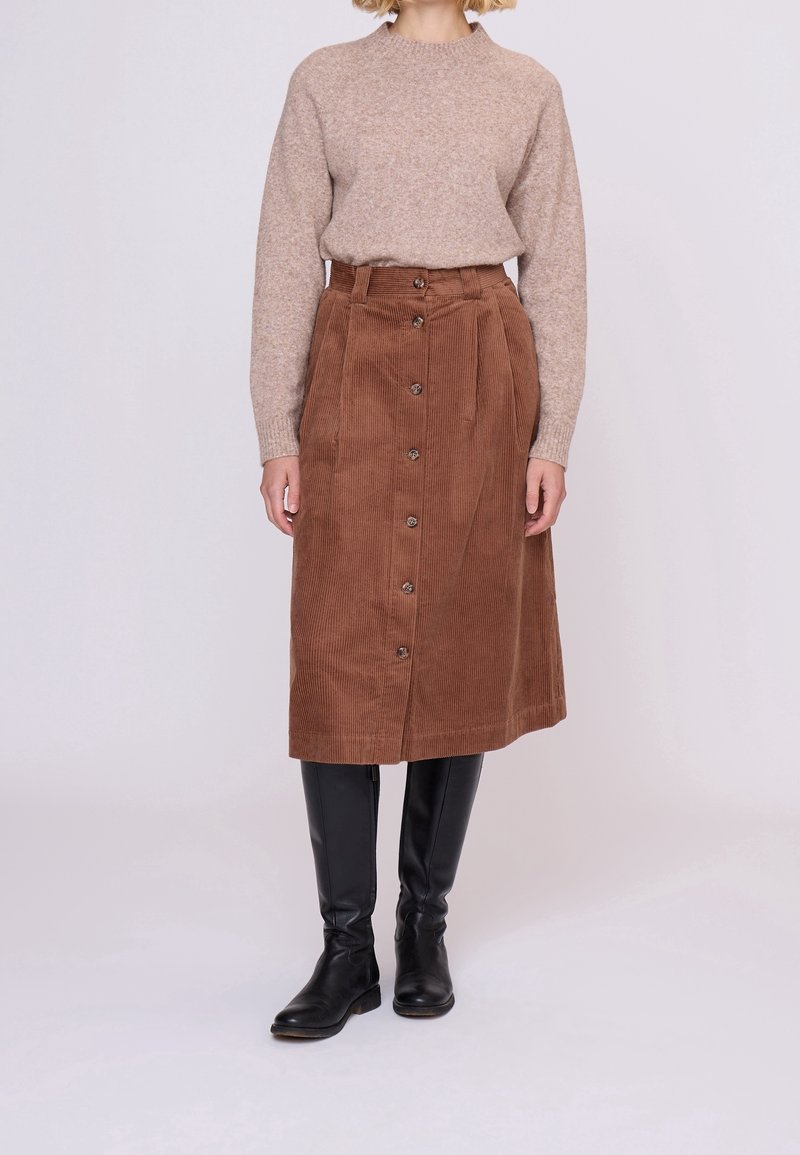 Femme portant un pull beige, une jupe midi en velours côtelé marron taille haute avec boutons, et des bottes noires montantes jusqu'aux genoux, debout devant un fond uni.