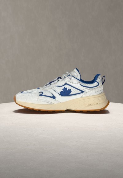 Baskets blanches avec un logo de feuille d'érable bleue, une doublure intérieure bleue et une semelle beige posées sur une surface claire sur un fond neutre.