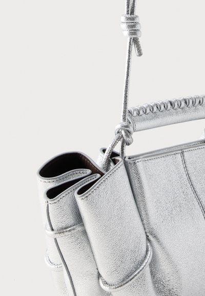 Κοντινή λήψη μιας ασημί δερμάτινης τσάντας με κομμένο λουρί και ραμμένη λεπτομέρεια στη λαβή, πάνω σε απλό φόντο.