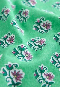 Grüner Stoff mit einem wiederholten Blumenmuster, das rosa Blumen und grüne Blätter zeigt, die auf einer strukturierten Oberfläche in dunklem Grün umrandet sind.