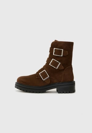 Bottines en daim marron avec trois accents de boucles en argent, un bout rond et une semelle en caoutchouc noir texturé pour une traction accrue.