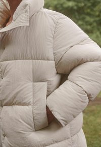 Lys beige pufferjakke med høj krave, kviltet tekstur, overdrevne ærmer og en forlomme. Har trykknapper til lukning.