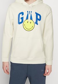 Krémová mikina s kapucí, přední kapsou, modrým textem "GAP" a grafikou žluté usměvavé tváře. Materiál z bavlněné směsi s kapucí na šňůrky.