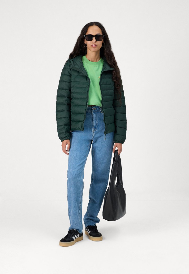 Groene pufferjack met gequilt textuur, gecombineerd met een felgroene t-shirt en blauwe jeans. Zwarte sneakers en een zwarte tas maken de outfit compleet.