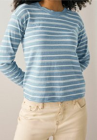 Lange mouwen, lichtblauwe gestreepte shirt met witte horizontale strepen; zachte textuur; ronde halslijn; gedragen met lichtbeige broek.