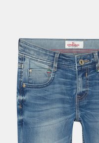 Niebieskie dżinsy z wyblakłym wykończeniem, pięcioma kieszeniami i mosiężnymi akcesoriami. Posiadają żółte przeszycia i białą metkę z czerwonym napisem.