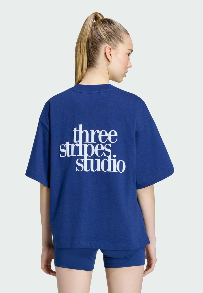 Femme aux cheveux blonds en queue de cheval porte un T-shirt bleu ample avec l'inscription « three stripes studio » et un short bleu assorti, vue de dos.
