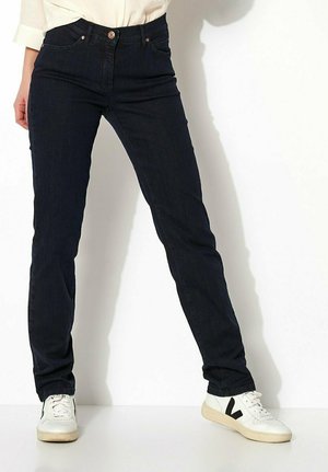 Person in dunkelblauen Jeans und weißen Sneakers mit schwarzem "V"-Logo, steht vor einem weißen Hintergrund mit leicht auseinanderstehenden Beinen.