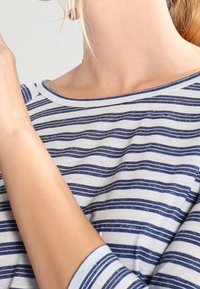 Närbild av en person som bär en vitt och marinblå randig skjorta med båturringning, vilket visar deras hals, axel och arm.