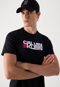 Fekete pamut póló, melynek elülső részén feltűnő fehér és piros "Calvin Klein" logó található. A modellen fekete sapka is van, fehér felirattal.