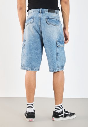 Homme portant un short cargo en jean bleu clair, des baskets noires et blanches et des chaussettes blanches à rayures noires, debout devant un fond uni.