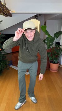 Grüner, flauschiger Hoodie, graue gestreifte Hose und silberne Turnschuhe. Hellfarbige Mütze mit Logo. Innenraum mit Pflanzen und festlicher Dekoration.