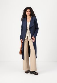 Navy trench coat with oversized collar, beige buttoned top, wide beige trousers, and brown handbag. Glossy black ankle boots complete the look.