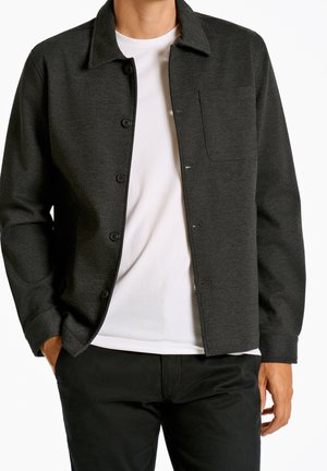 Homme portant une veste boutonnée gris foncé par-dessus un t-shirt blanc et un pantalon noir, une main dans la poche, sur un fond blanc uni.