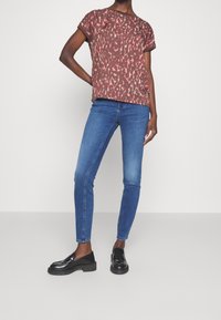 Persona con una blusa abstracta de manga corta en tonos rojos y marrones, jeans ajustados azules y mocasines negros, de pie con una mano en la cadera contra un fondo blanco.