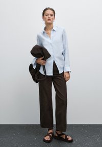 Camicia button-up in lino azzurro chiaro, pantaloni ampi in suede marrone e sandali neri con cinturini, con una borsa marrone scuro appoggiata su un braccio.