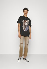 Svart bomull T-skjorte med grafisk design som viser en hånd og en sommerfugl. Kombinert med lysebrune ripped jeans og svarte sneakers.