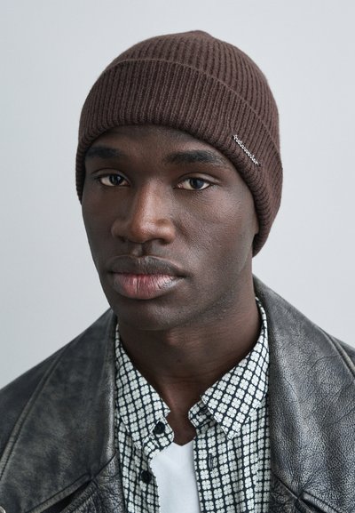 Brązowa prążkowana dzianinowa czapka z małym logo, noszona z czarną skórzaną kurtką i wzorzystą koszulą. Neutralne tło.