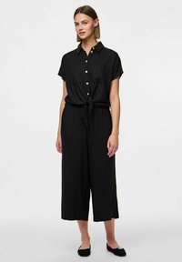 Črn laneni kombinezon s ovratnikom, kratkimi rokavi, sprednjimi gumbi in pasom na vezanje. Ima široke hlače in lahkotno teksturo.