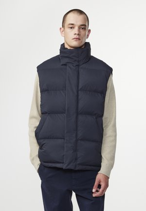 Navy puffer vest with high collar, quilted design, front zipper closure, and no sleeves, worn over a beige sweater. Smooth texture.