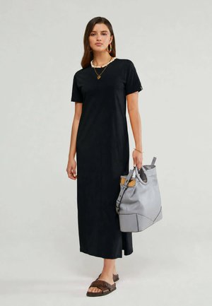 Donna che indossa un maxi abito nero a maniche corte, con una grande borsa grigia, accessoriata con collane a strati, orecchini colorati e sandali marroni.