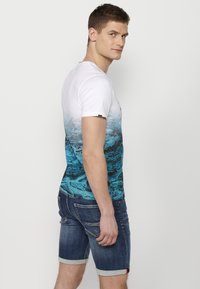 Graphic t-shirt featuring a white top and a blue ocean wave pattern on the lower half. Paired with denim shorts, showing rolled cuffs.