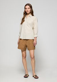 Jasnobeżowa koszula zapinana na guziki z podwiniętymi rękawami, w połączeniu z brązowymi szortami cargo z dwoma kieszeniami. Uzupełniona klapkami.