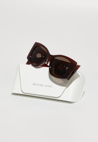 Rjave prevelike sončne očala z temnimi lečami, postavljene na belo teksturirano etuije z embosiranim logotipom "MICHAEL KORS".