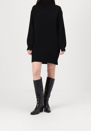 Femme portant une robe-pull à col roulé côtelée noire associée à des bottes en cuir noir brillant montant jusqu'au genou avec un talon bloc.