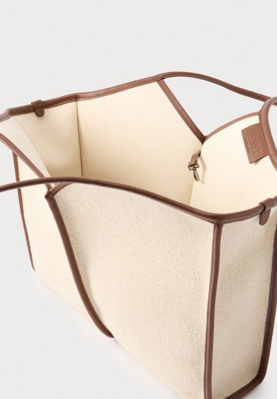 Sac fourre-tout en toile beige avec garniture et anses en cuir marron, ouverture supérieure, et petite poche intérieure zippée avec languette en cuir.