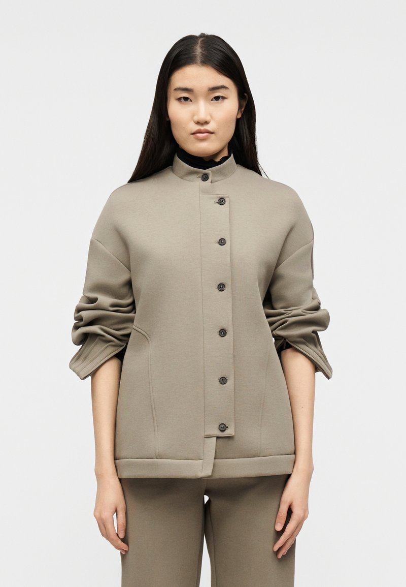 Γυναίκα με μακριά σκούρα μαλλιά που φοράει ταμπά σακάκι με κουμπιά και τυλιγμένα μανίκια, καθώς και ταιριαστό παντελόνι, στέκεται μπροστά σε απλό φόντο.