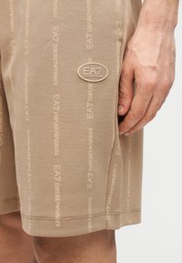 Švelnios medžiagos smėlio spalvos šortai su pasikartojančiu logotipo raštu ir apvalia logotipo aplikacija ant šlaunies. Apačia šiek tiek susukta.