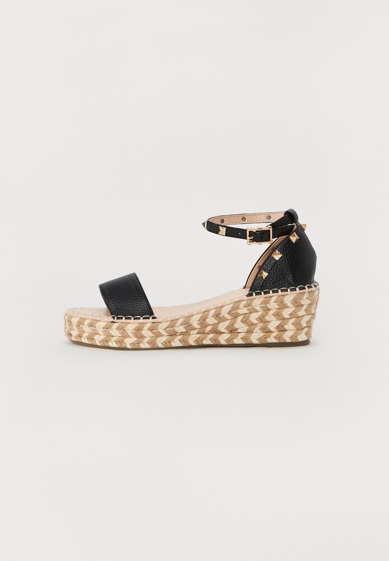 Sandale compensée en cuir noir avec semelle plateforme espadrille tissée, bride cheville avec boucle dorée et clous dorés au talon.