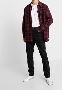 Hombre con una camisa de cuadro rojo y negro sobre una camiseta blanca, jeans negros y zapatillas blancas, de pie con una mano en el bolsillo.