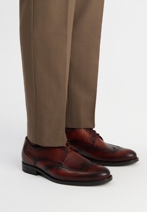 Zapatos de vestir de cuero marrón con detalles perforados, combinados con pantalones beige, que muestran un diseño clásico de cordones y un brillo sutil.