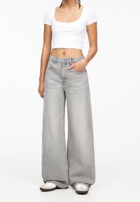 Donna che indossa un top bianco a maniche corte con scollatura a giro, jeans grigi a vita alta e gamba larga, e sneakers bianche, in piedi con una mano in tasca.