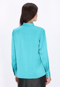 Turkusowa bluzka z długim rękawem i wysokim kołnierzem, o gładkiej satynowej fakturze i guzikach przy mankietach. Posiada delikatne fałdy na ramionach.
