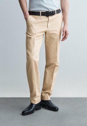 Homme portant un pantalon beige, une ceinture noire, une chemise bleu clair et des chaussures en cuir noir, debout sur un sol gris devant un fond gris clair.